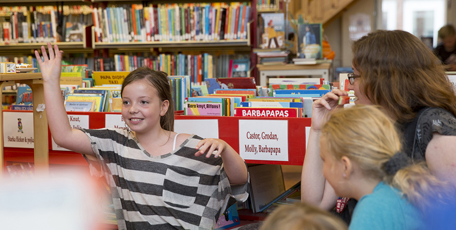 barngrupp på biblioteket. Glad tjej håller upp handen. 