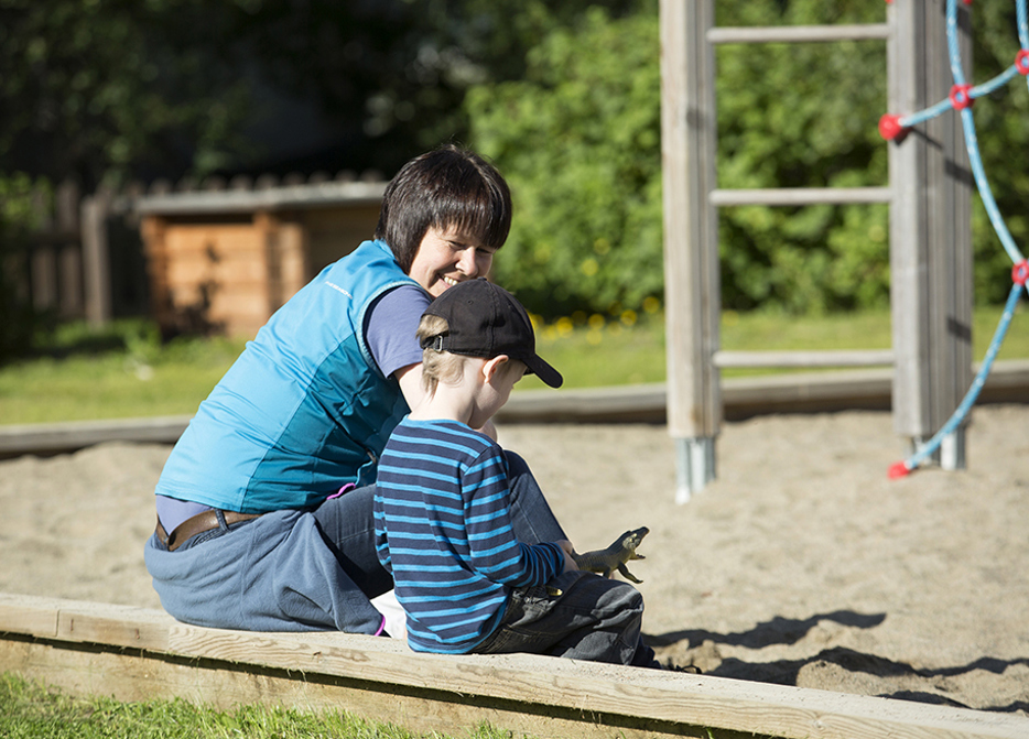 Kvinnlig förskolefröken sitter med pojke på kanten av sandlådan och pratar. Solig dag.