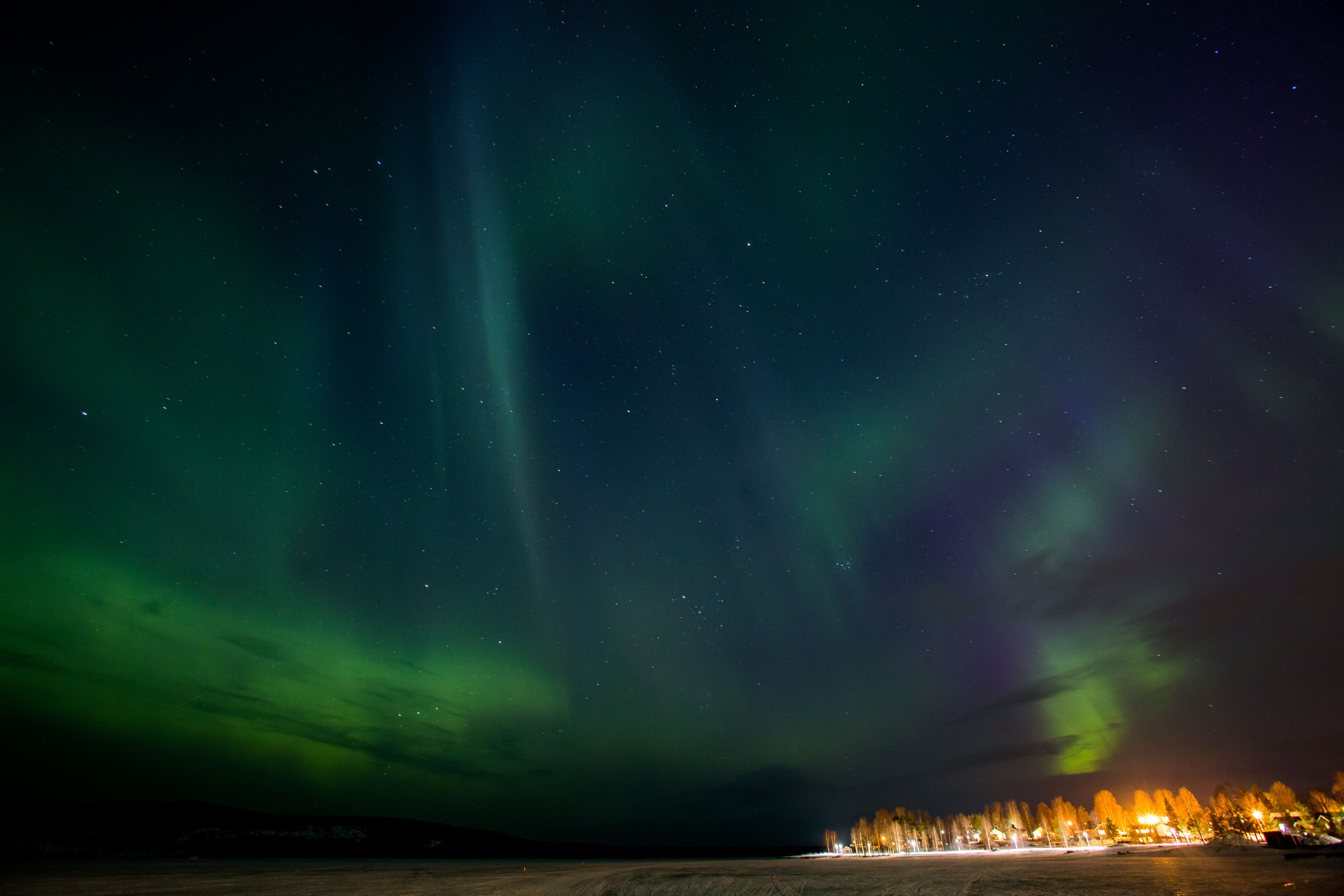 Vy över vinterlandskap. Lampor från bostäder lyser upp den mörka vinternatten som även pryds av ett grönt norrsken. 